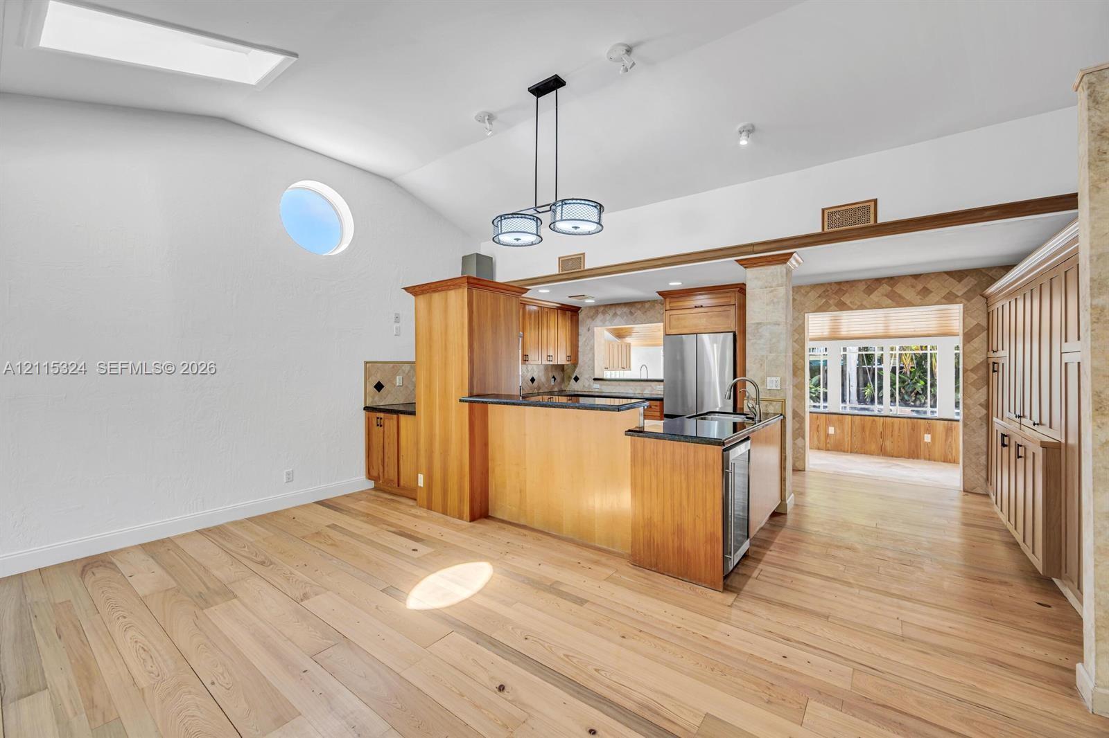 Vaulted ceilings featuring a skylight and circular accent window, wood-finish flooring, custom cabinetry with dark countertops, and a modern multi-pendant light fixture