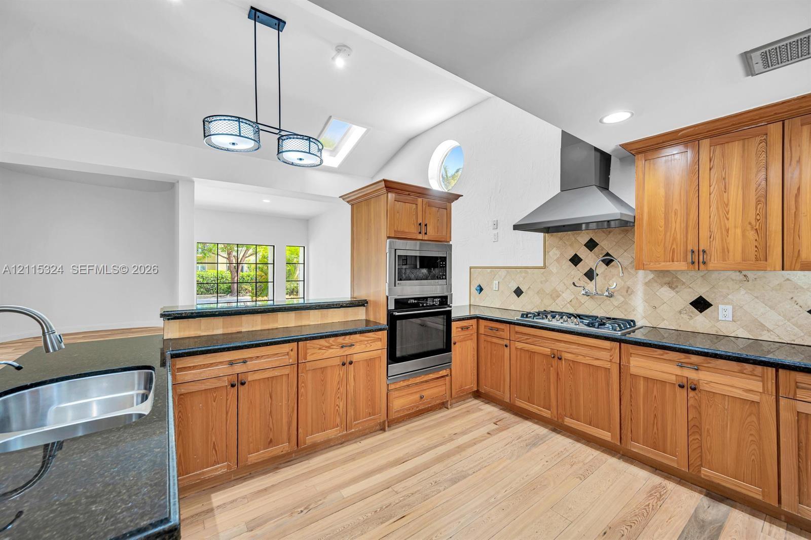 Kitchen featuring wood cabinetry, dark stone countertops, stainless steel appliances, a tiled backsplash with diamond accents, and a vaulted ceiling with skylights