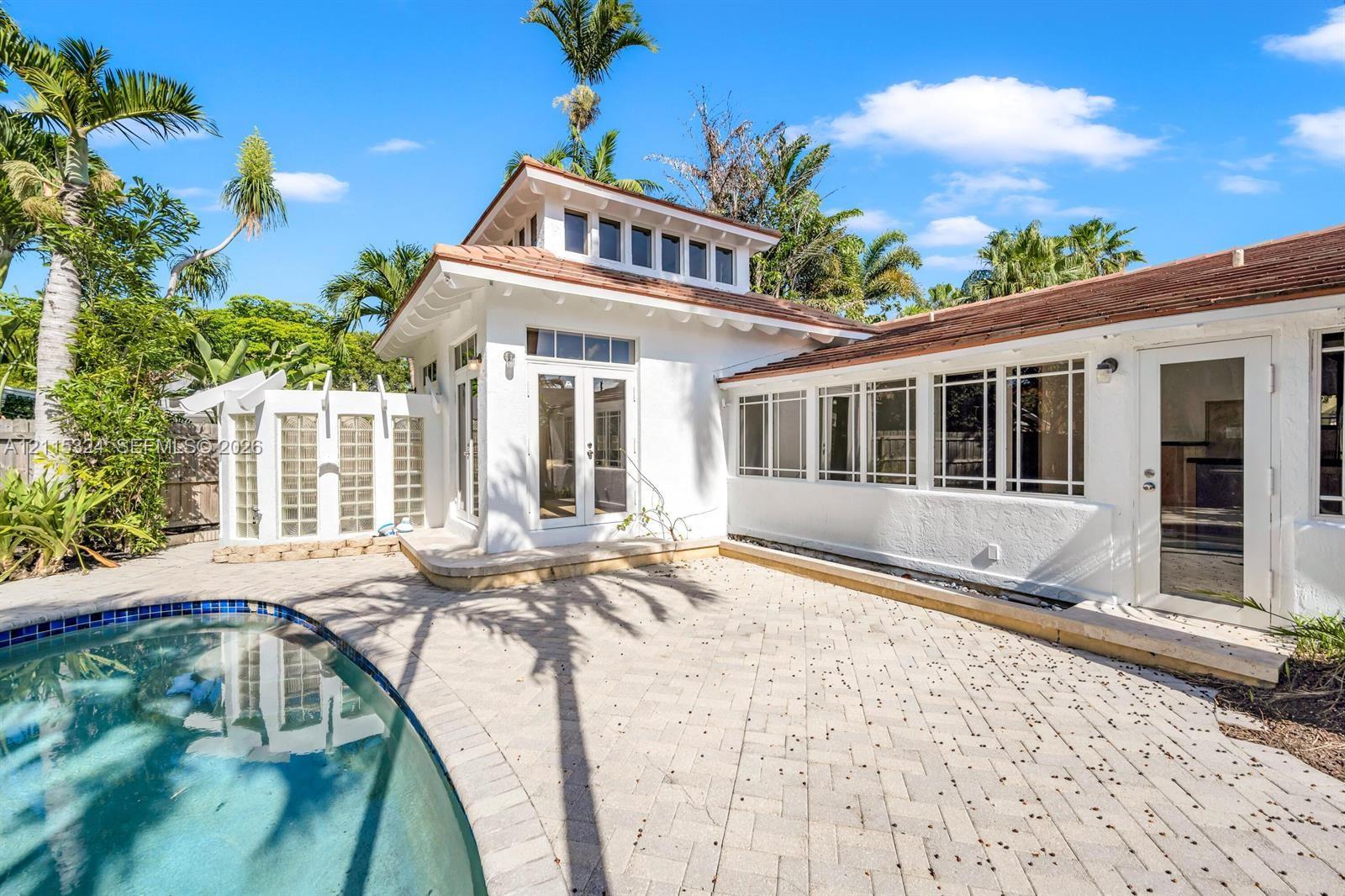 Exterior featuring white stucco siding, a curved in-ground pool, expansive stone-paver patio, glass-block privacy wall, and multi-pane transom windows