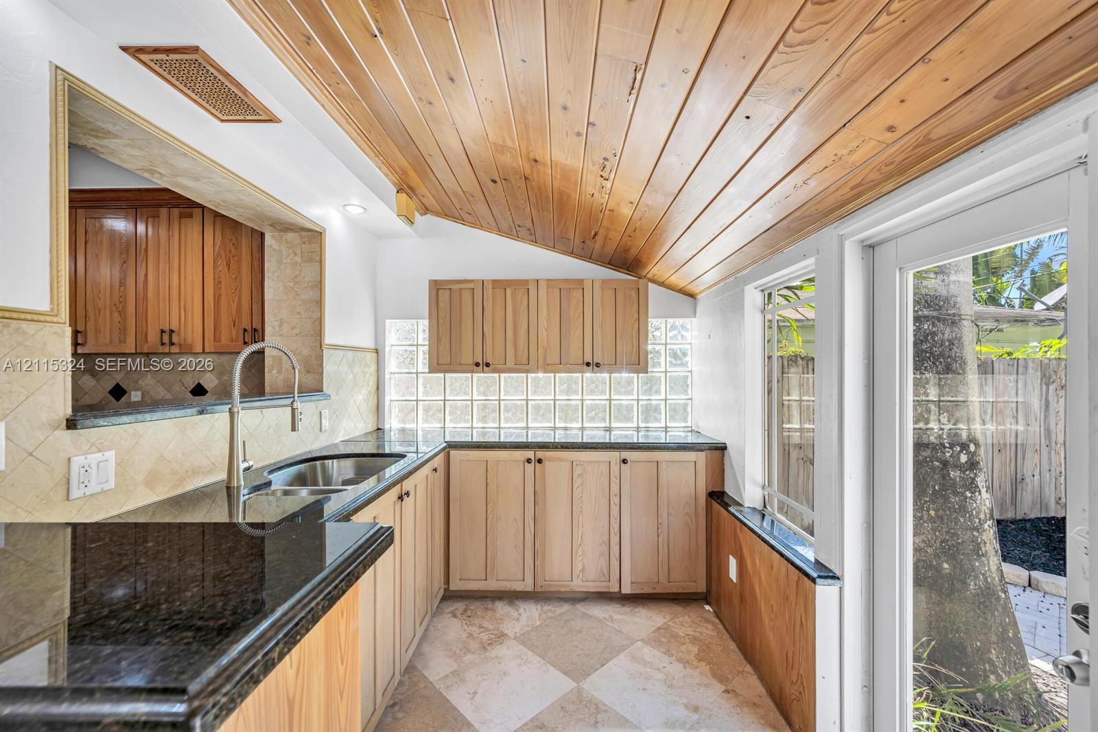 Vaulted tongue-and-groove wood ceiling, polished stone countertops, light wood cabinetry, glass block accent wall, and stone-tile flooring