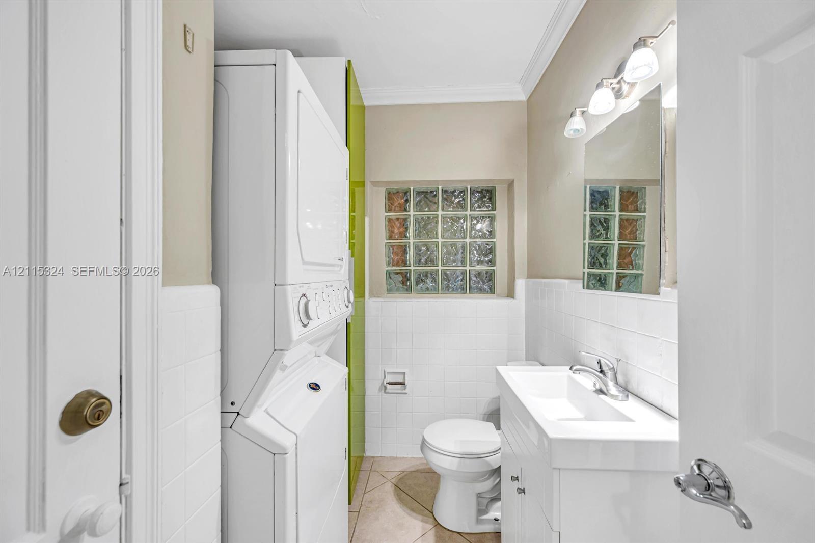 Combination laundry area and bathroom featuring a stacked washer and dryer, square glass block window, white vanity with integrated sink, and polished chrome fixtures