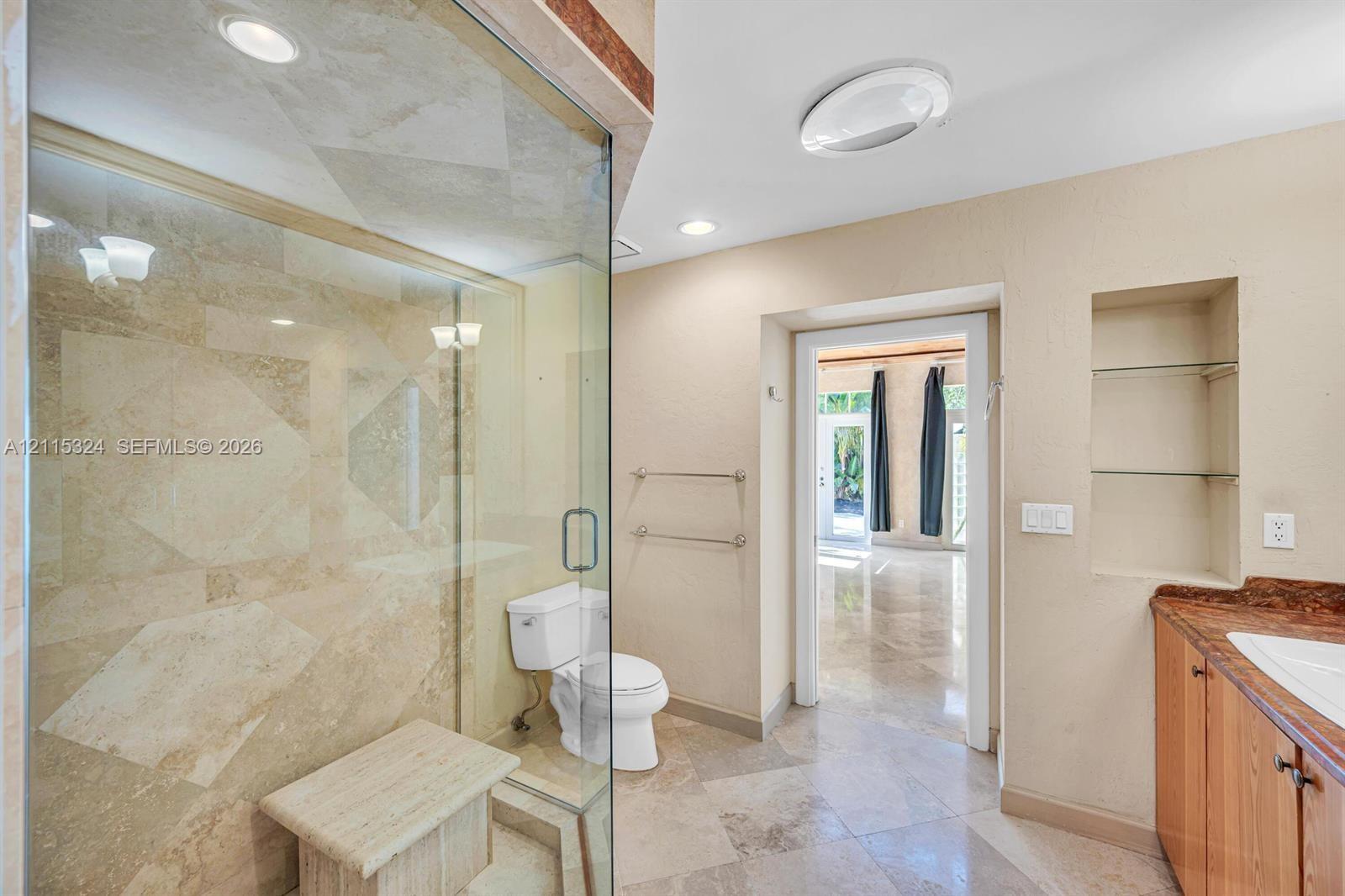 Walk-in shower with natural stone tile surround and built-in bench, glass enclosure, stone tile flooring, built-in wall shelving, and cabinetry with stone countertop