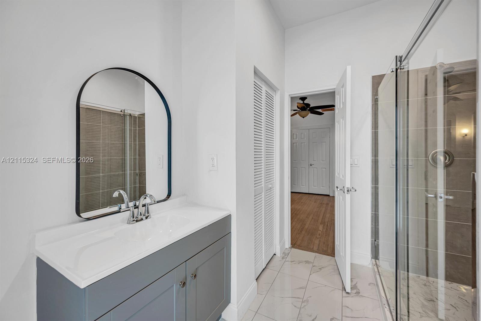 Bathroom featuring a grey vanity with a rectangular sink, arched mirror, glass-enclosed shower with tile surround, and tile flooring