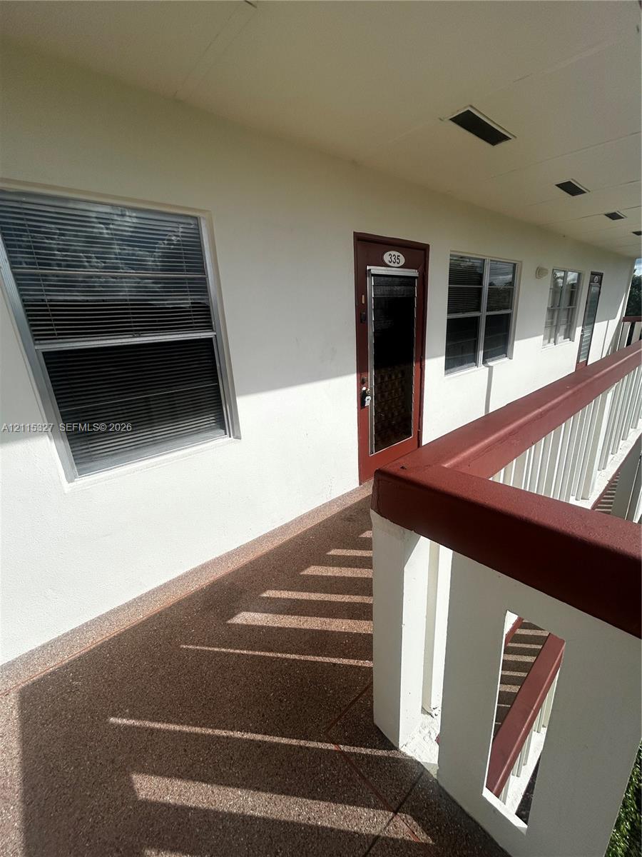 Exterior walkway featuring textured stone-aggregate flooring, a white railing with a dark-tone handrail, and multiple windows with integrated blinds