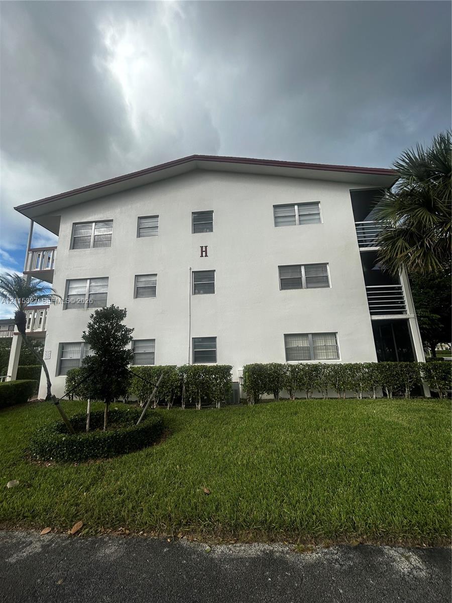 Three-story exterior featuring a light-toned facade, horizontal sliding windows, covered balconies with railings, and manicured shrubbery