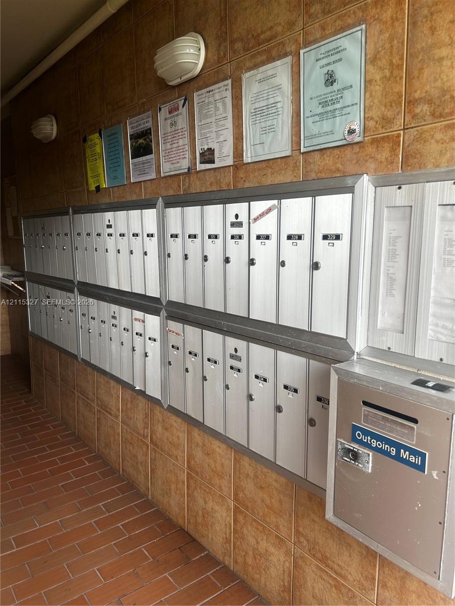 Wall-mounted metallic mailbox station featuring individual locked compartments and an integrated outgoing mail slot, set against a ceramic-tiled wall with attached overhead lighting