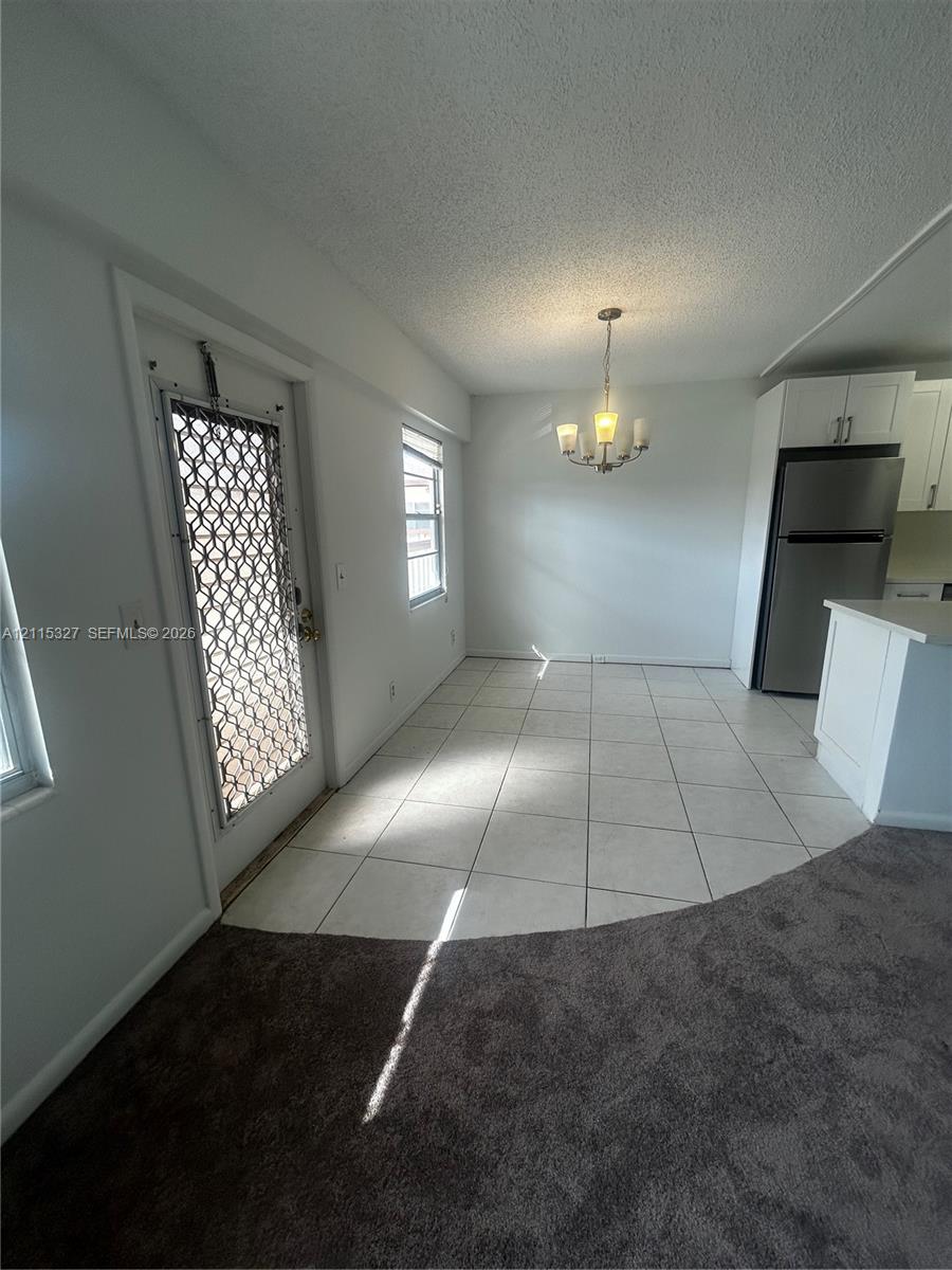 Neutral-toned ceramic tile flooring, white shaker-style cabinetry, stainless steel refrigerator, and multi-arm chandelier lighting