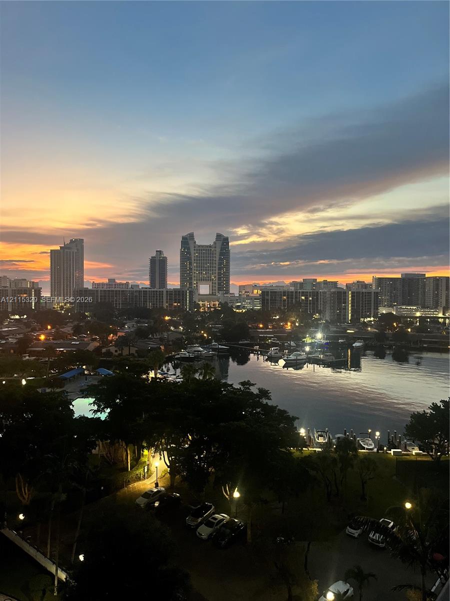 Panoramic cityscape featuring high-rise buildings, a waterfront marina, and mature landscaping under a sunset sky