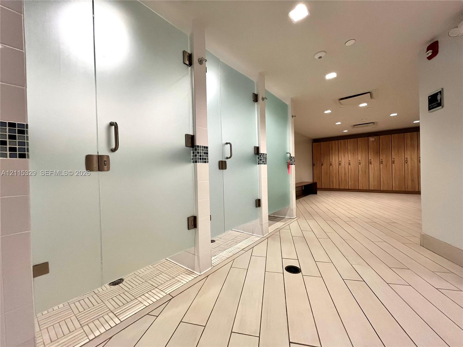 Frosted glass shower stalls with brushed metal hardware, mosaic tile flooring, wood-finish lockers, and recessed lighting