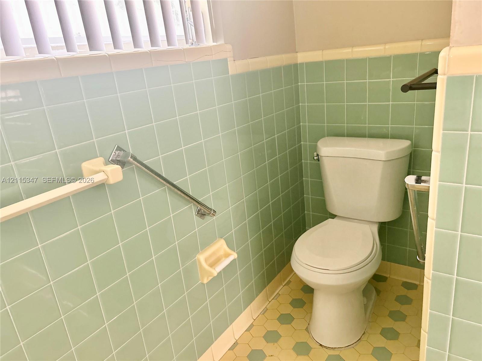 Restroom featuring light green square wall tiling, cream-colored trim, hexagon-patterned flooring, a two-piece toilet, and metallic wall-mounted hardware