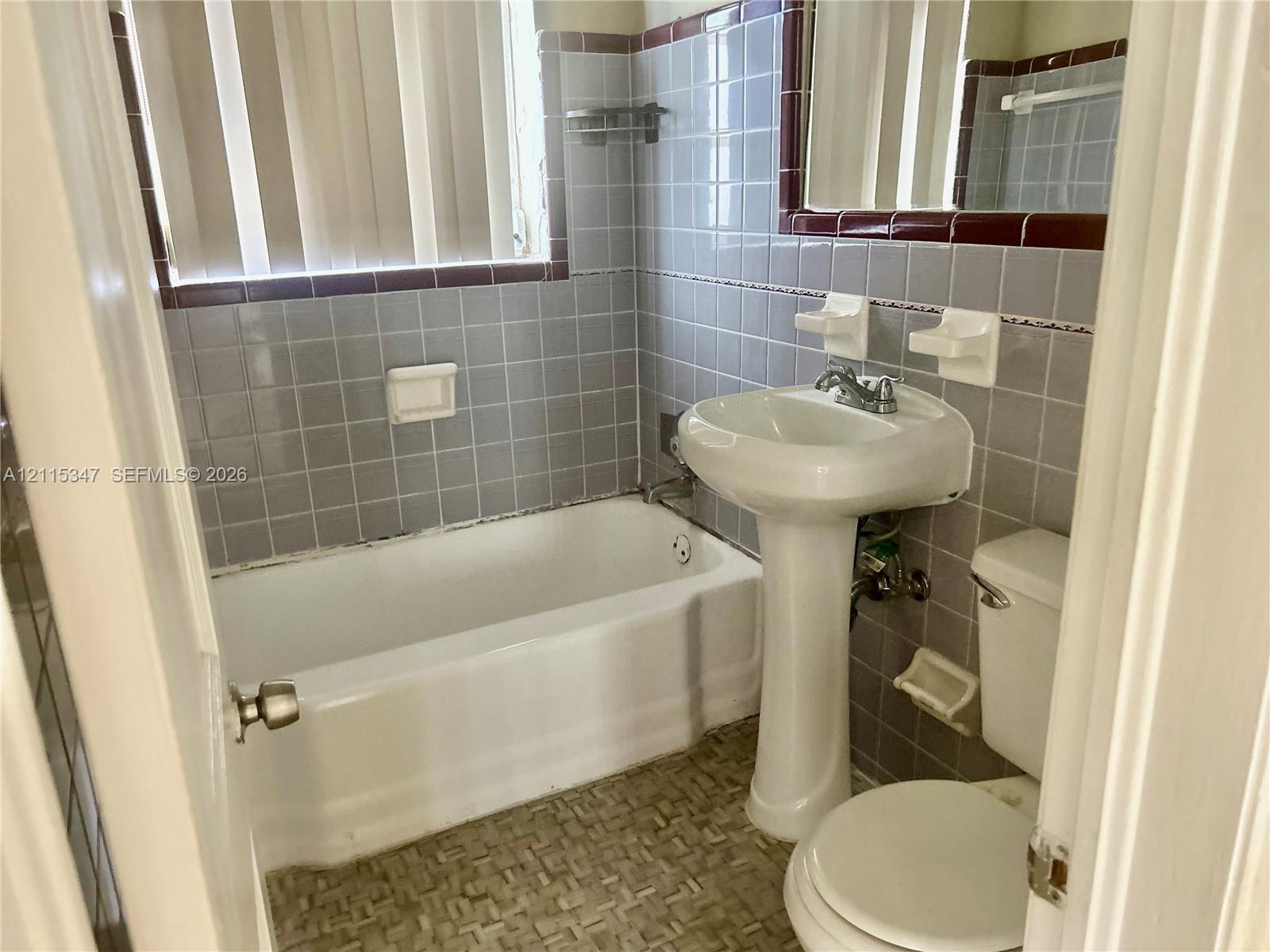 Bathroom featuring a white pedestal sink, a standard tub with shower head, patterned tile flooring, and light-toned wall tile with dark decorative trim