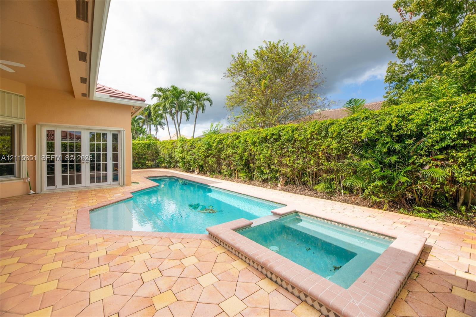 In-ground swimming pool and attached spa with a paved stone deck, covered patio, and perimeter privacy hedging