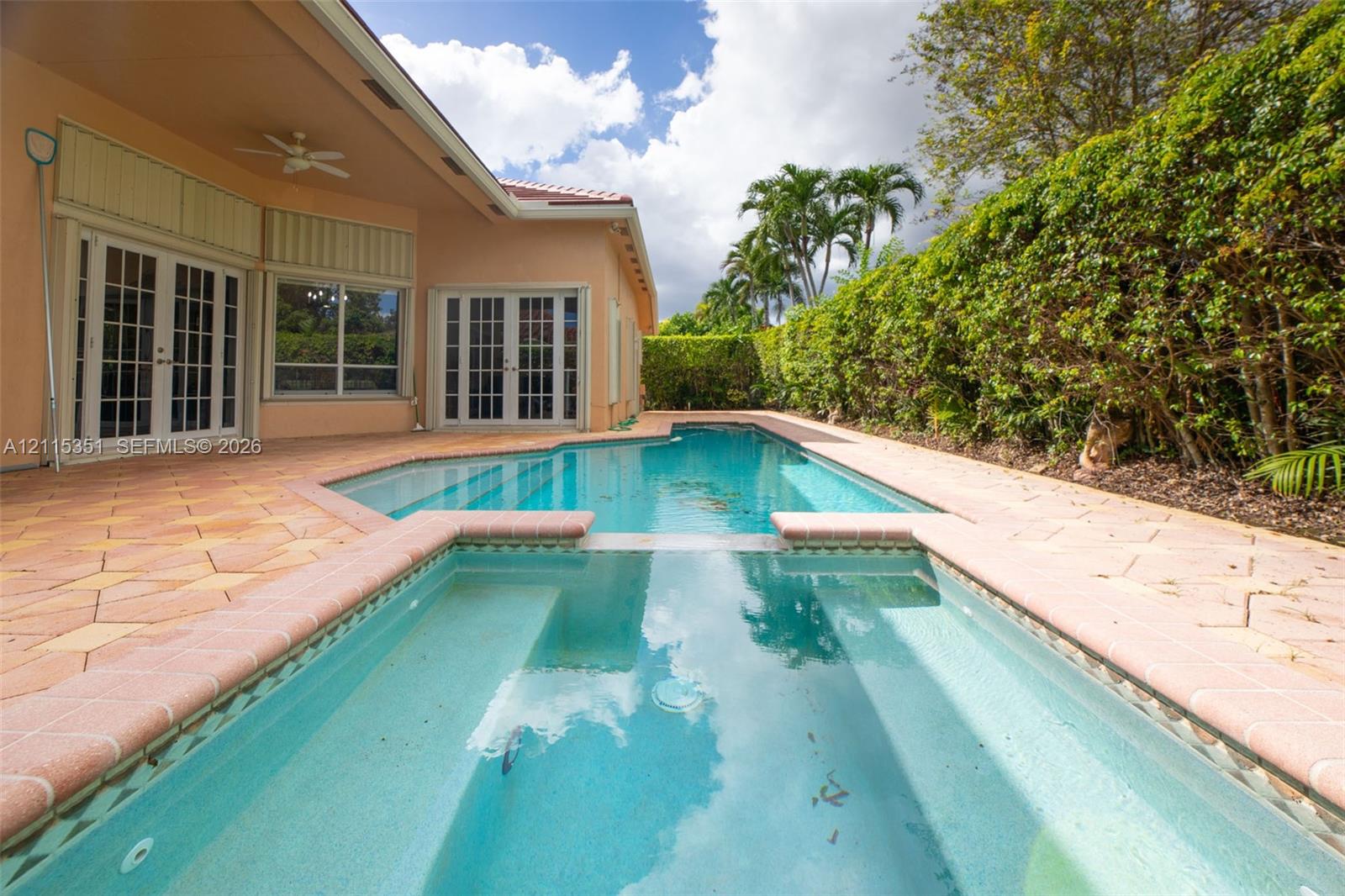 In-ground swimming pool with integrated spa, paver patio, covered porch with ceiling fan, and surrounding privacy hedging