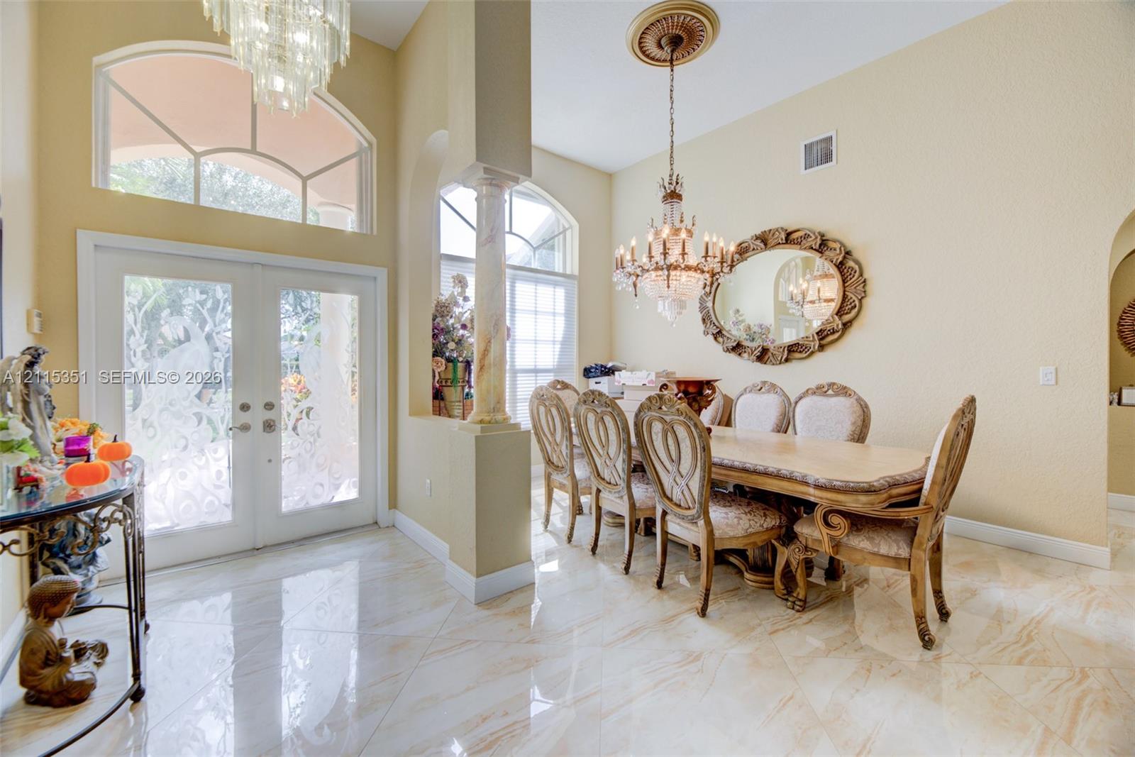 Foyer and dining space featuring large-format tile flooring, dual glass-paneled entry doors with arched transom windows, a decorative column, and dual crystal chandeliers Foyer and dining space featuring large-format tile flooring, dual glass-paneled entry doors with arched transom windows, a decorative column, and dual crystal chandeliers