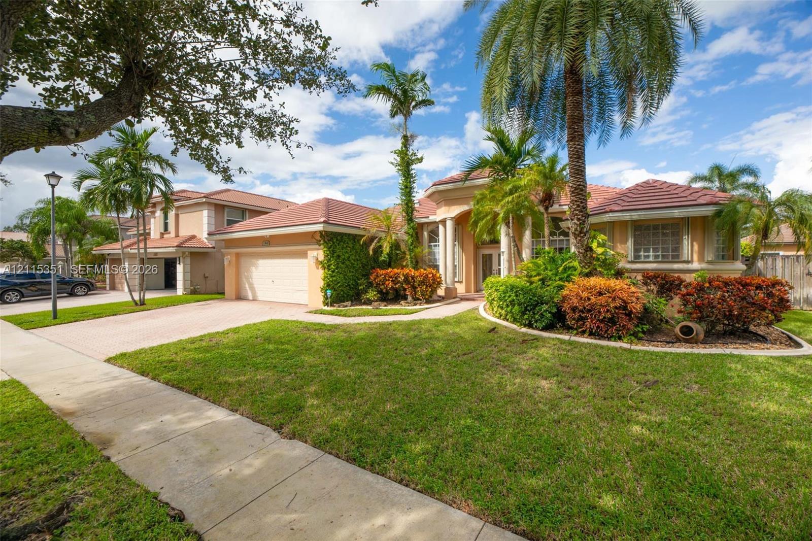 Stucco exterior residence featuring a multi-hip tile roof, paved driveway, column-accented entryway, and mature tropical landscaping