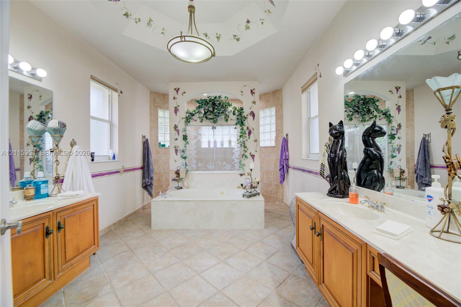Bathroom featuring dual wood-finish vanities with solid-surface countertops, a central jetted tub with tiled surround, glass block windows, and a coffered ceiling with a central light fixture