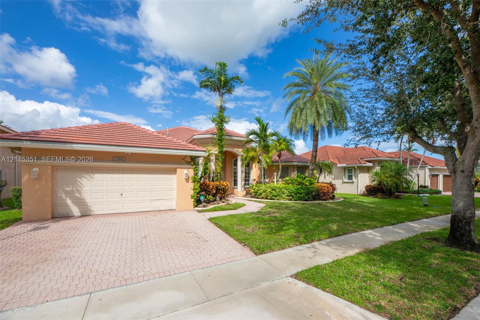 Stucco exterior featuring a red tile roof, attached two-car garage, brick paver driveway, and mature landscaping