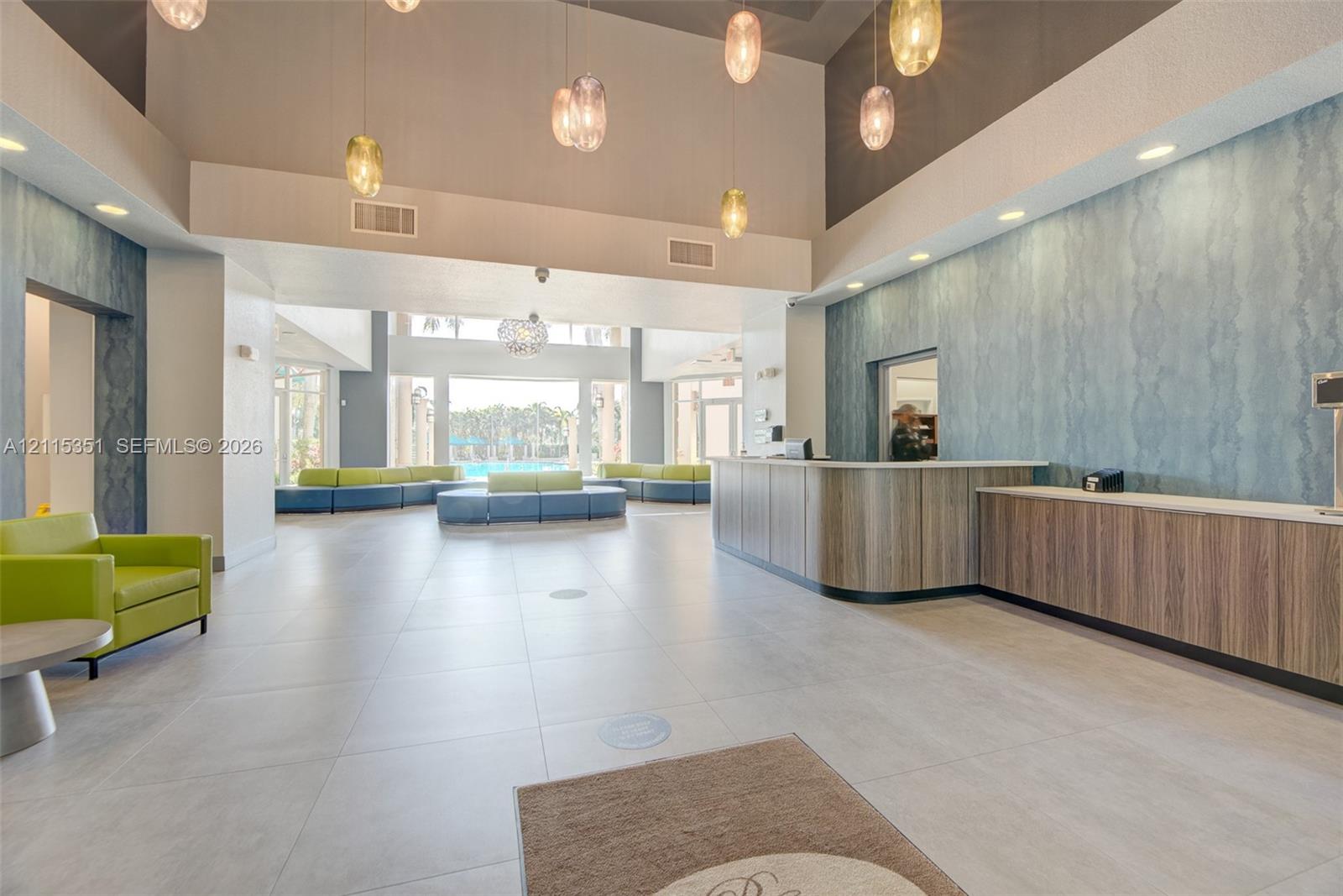 Grand lobby featuring high ceilings, large-format tile flooring, textured accent wall, integrated custom reception desk, and multi-tier pendant lighting