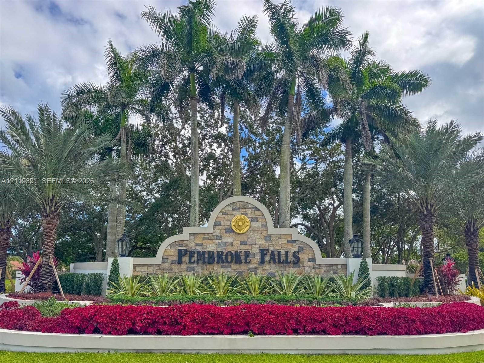 Community entrance signage featuring masonry stonework, dual exterior light fixtures, mature palm trees, and tiered landscaping
