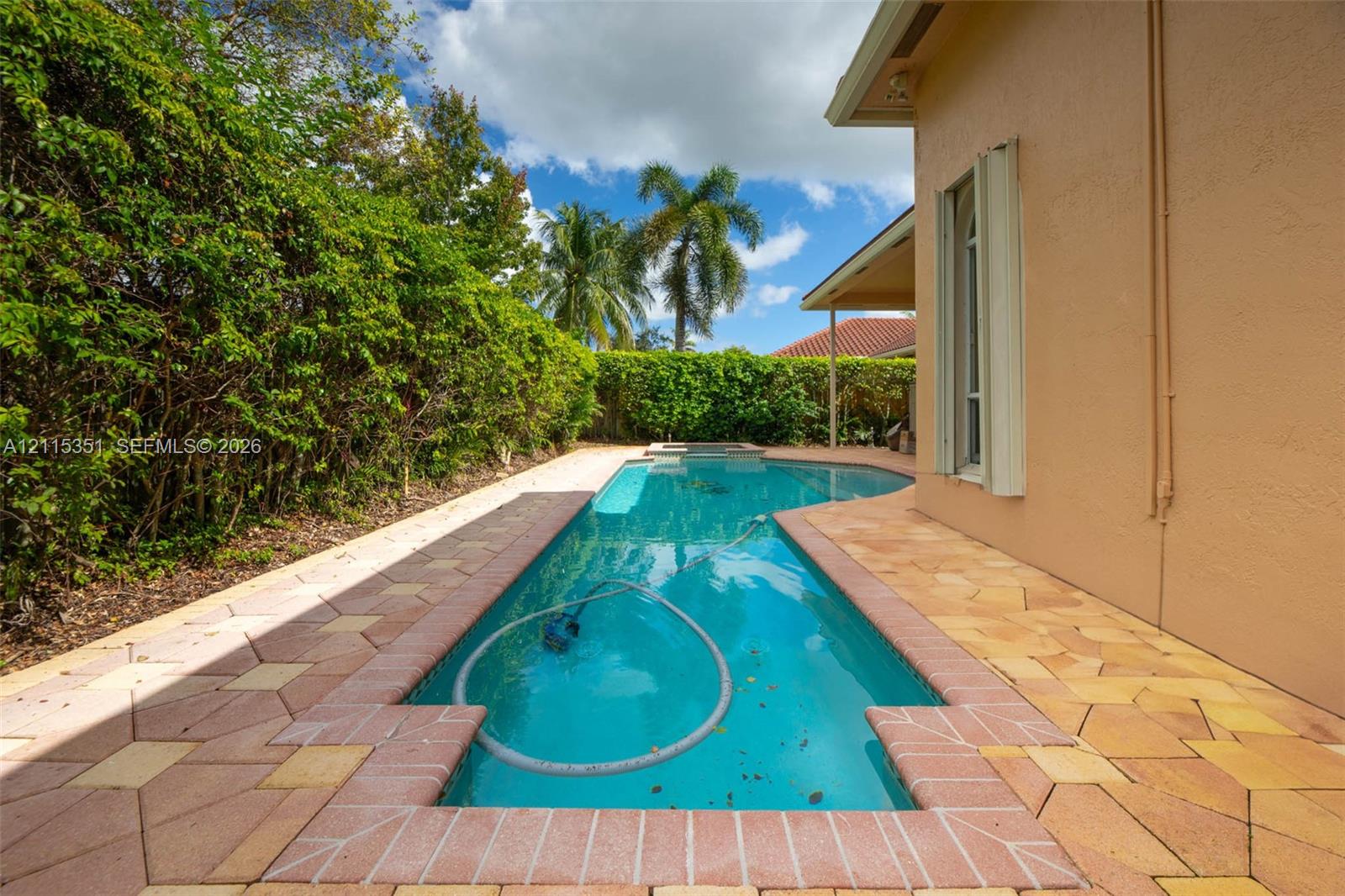 In-ground swimming pool surrounded by a multi-tonal paver deck, accented by mature perimeter hedge landscaping and a covered patio area