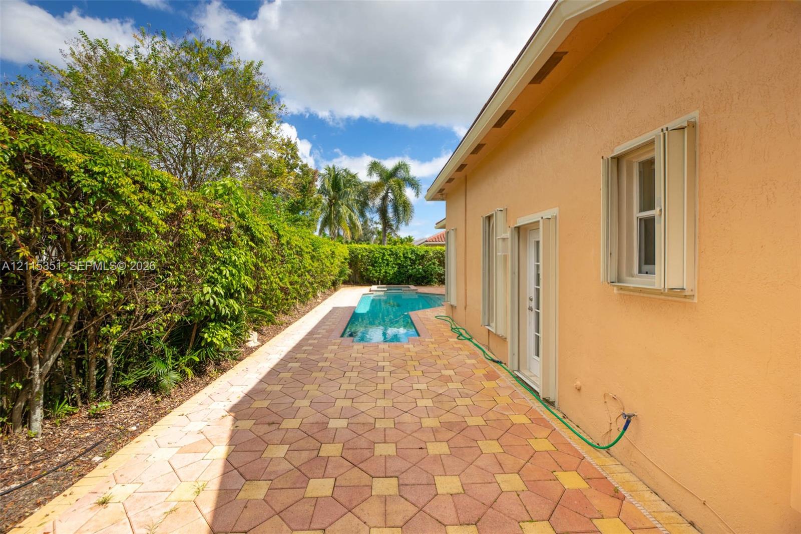 Outdoor living area featuring patterned paver hardscaping, a geometric swimming pool, exterior wall shutters, and manicured hedge landscaping