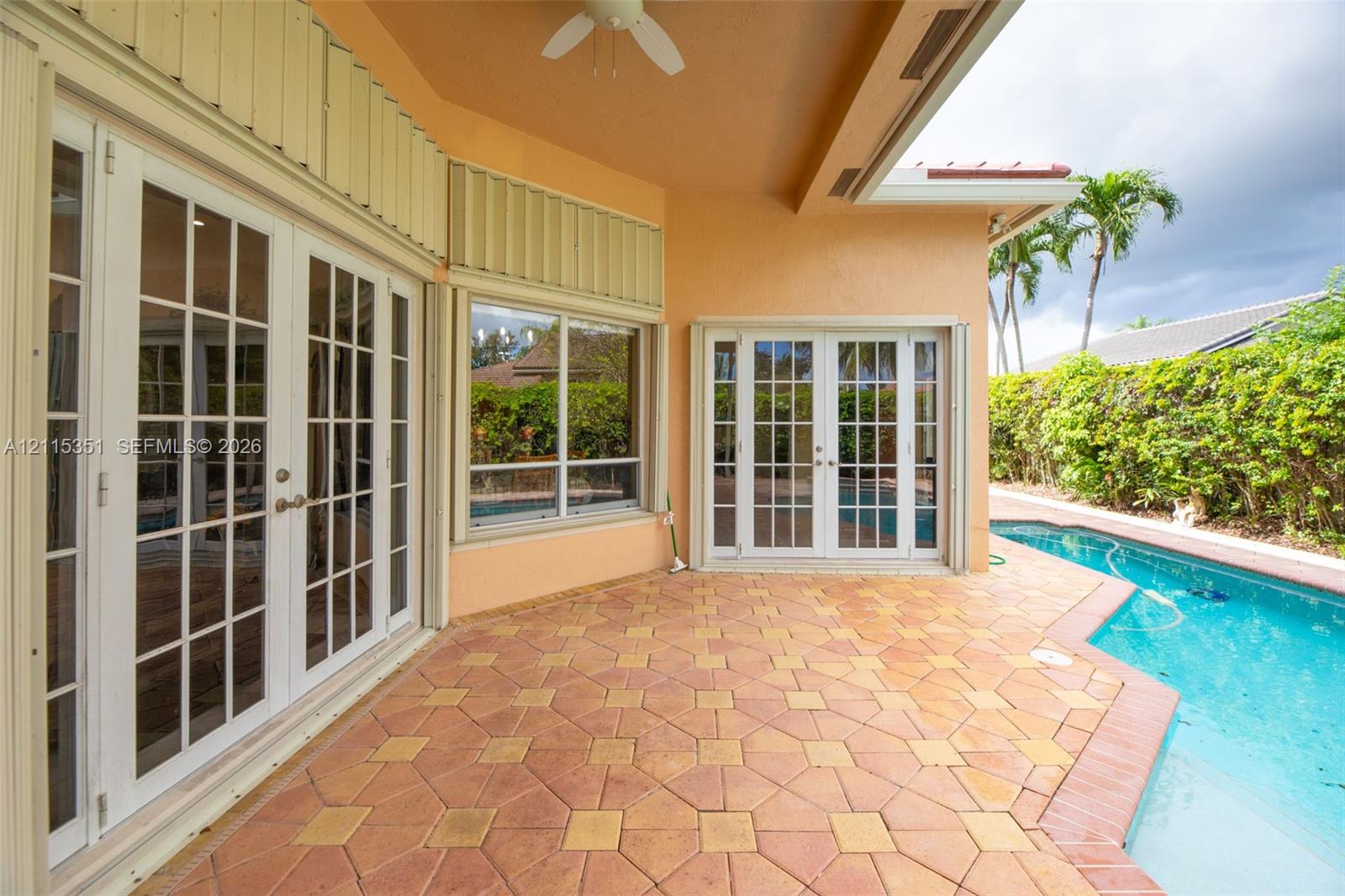 Covered patio featuring patterned stone pavers, multiple sets of white French doors, integrated ceiling fan, and an in-ground swimming pool