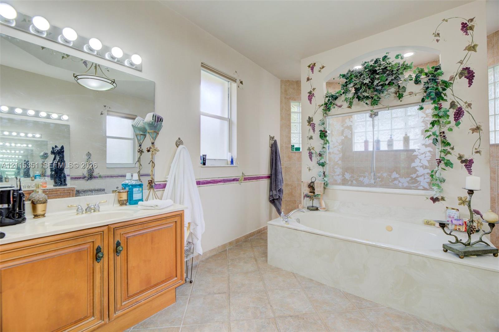Bathroom featuring a wood-finish vanity with a molded countertop, expansive wall-mounted mirror with integrated lighting, oversized soaking tub with an arched glass-block surround, and tiled flooring