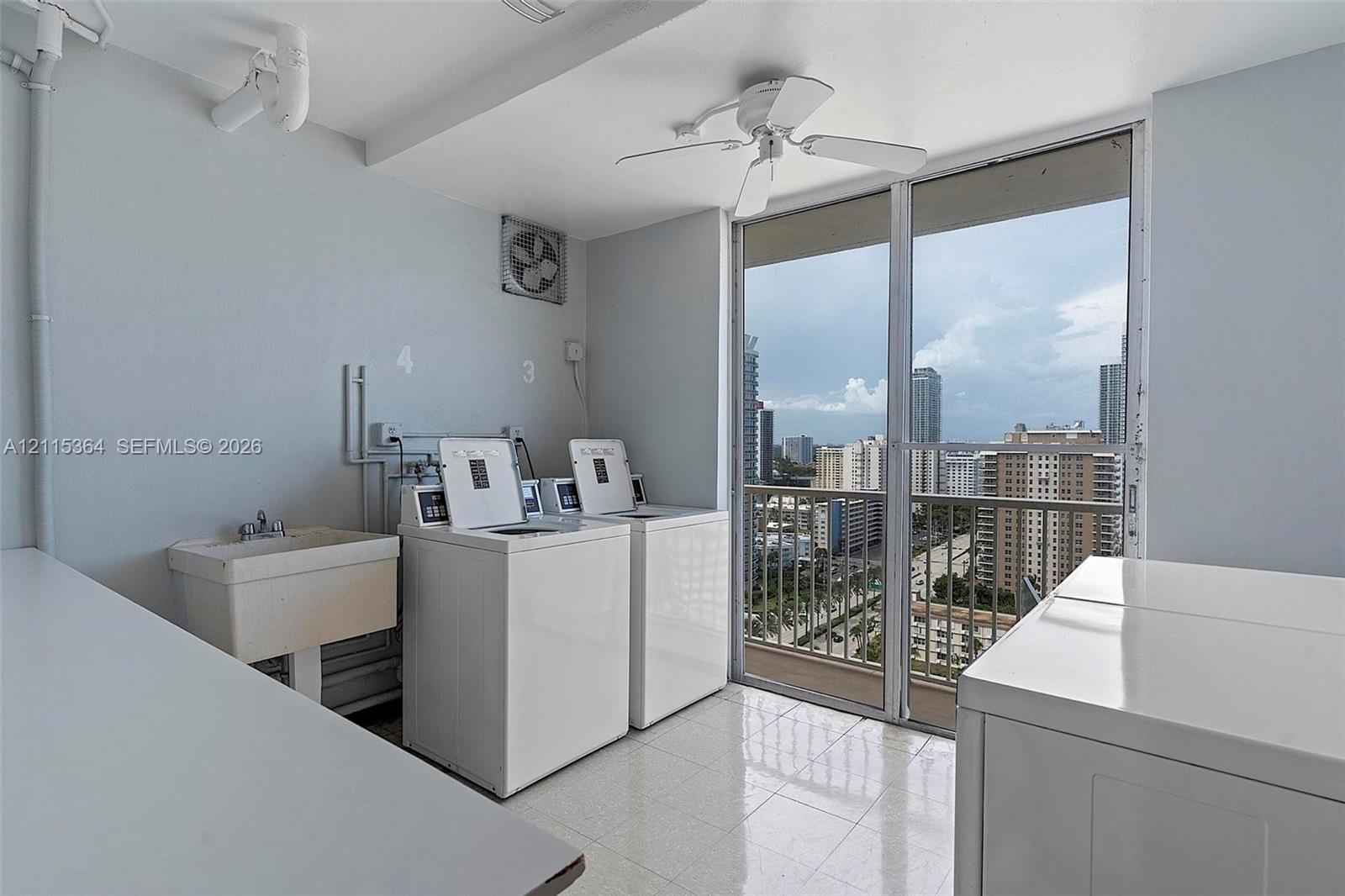 Laundry area featuring tile flooring, a wall-mounted utility sink, ceiling-mounted fan, and floor-to-ceiling glass sliding doors opening to an exterior balcony