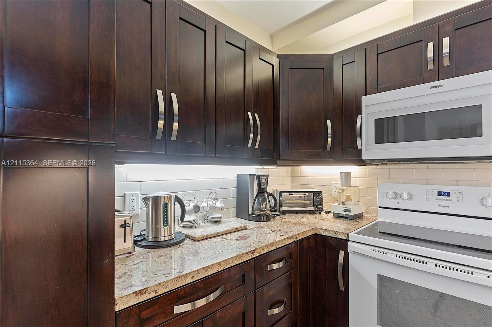 Dark-stained wood cabinetry, stone countertops, integrated under-cabinet lighting, subway tile backsplash, and white appliance suite