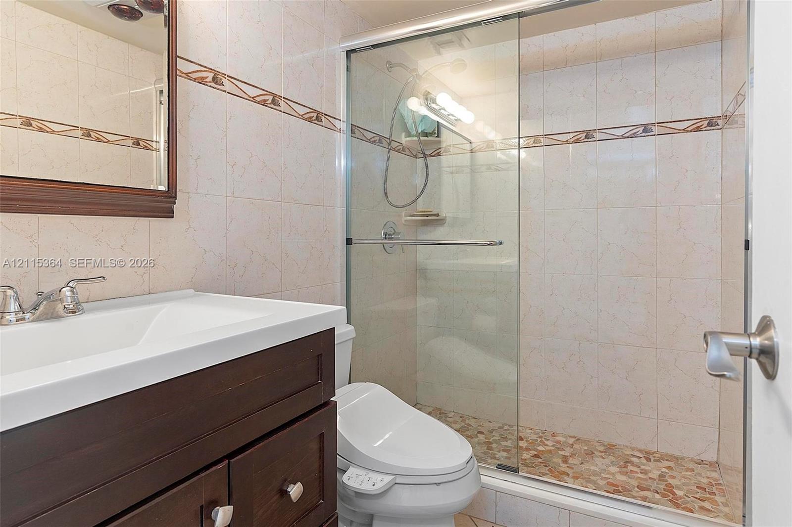 Bathroom featuring a dark wood-finish vanity with a white integrated basin, tile wall cladding with a decorative border, a glass-enclosed shower with pebble-stone flooring, and a wall-mounted mirror with a dark wood frame
