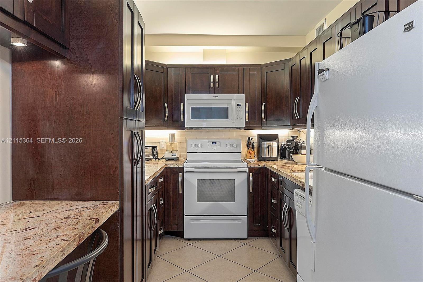 Dark-stained wood cabinetry, stone countertops, under-cabinet lighting, and white appliances including an electric range and microwave