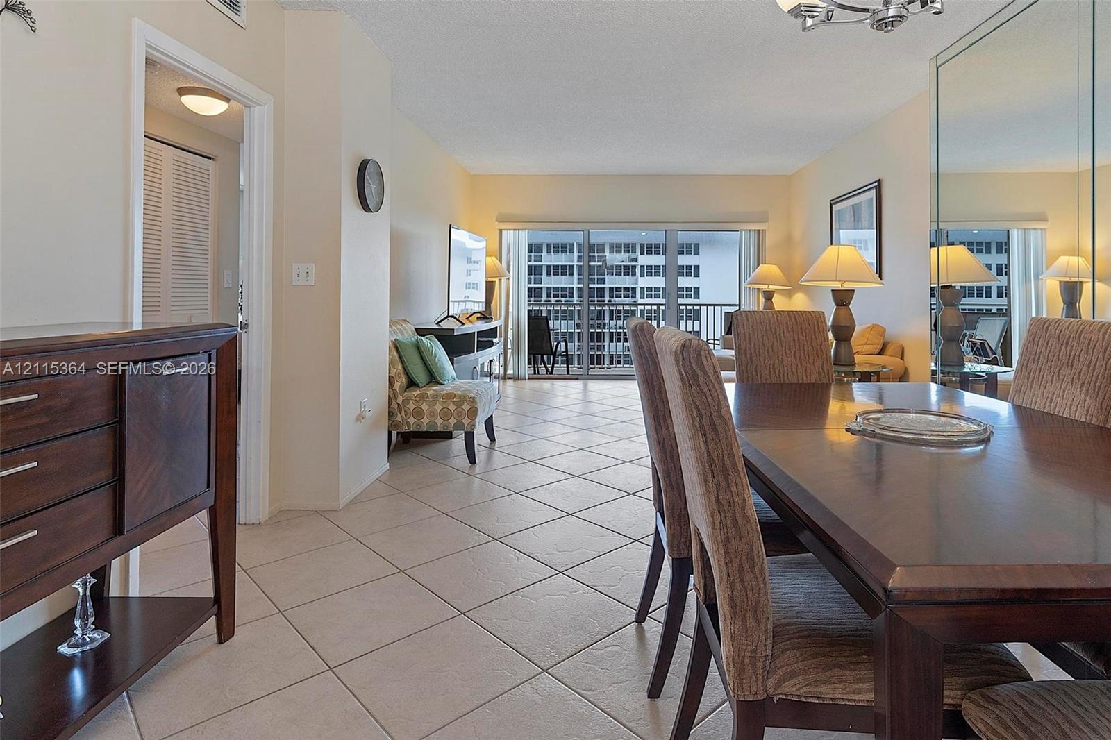 Open-concept living space featuring neutral ceramic tile flooring, floor-to-ceiling mirrored wall panels, sliding glass balcony doors, and recessed ceiling lighting