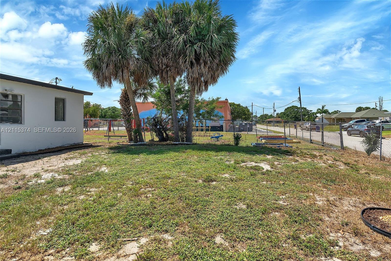 Exterior yard space featuring mature palm trees, perimeter chain-link fencing, and a combination of grass and sand landscaping