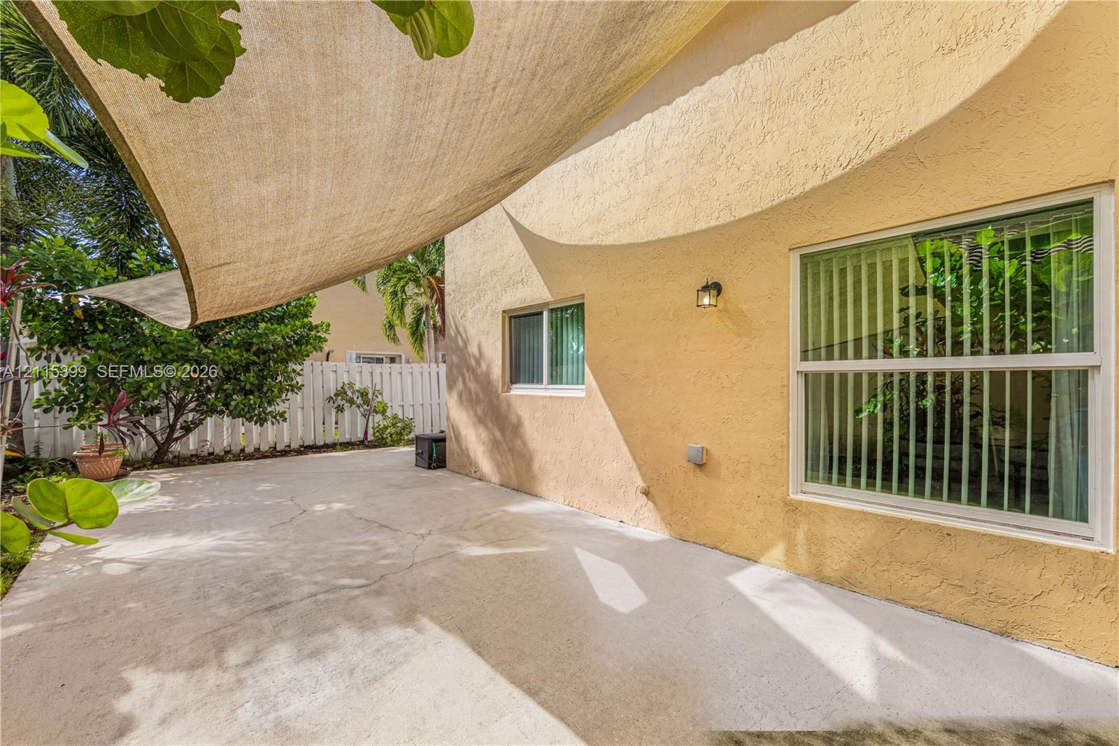 Stucco patio area featuring a tension shade sail, concrete flooring, exterior wall-mounted lighting, and a white privacy fence