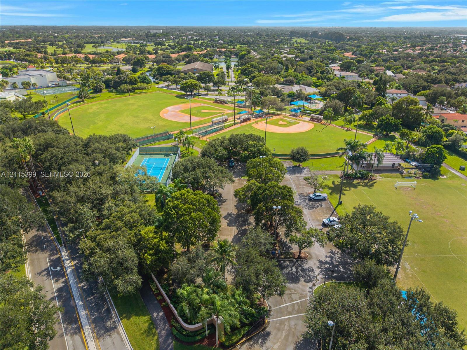Aerial perspective of community recreation grounds featuring baseball diamonds, a tennis court, expansive grassy fields, and paved parking areas