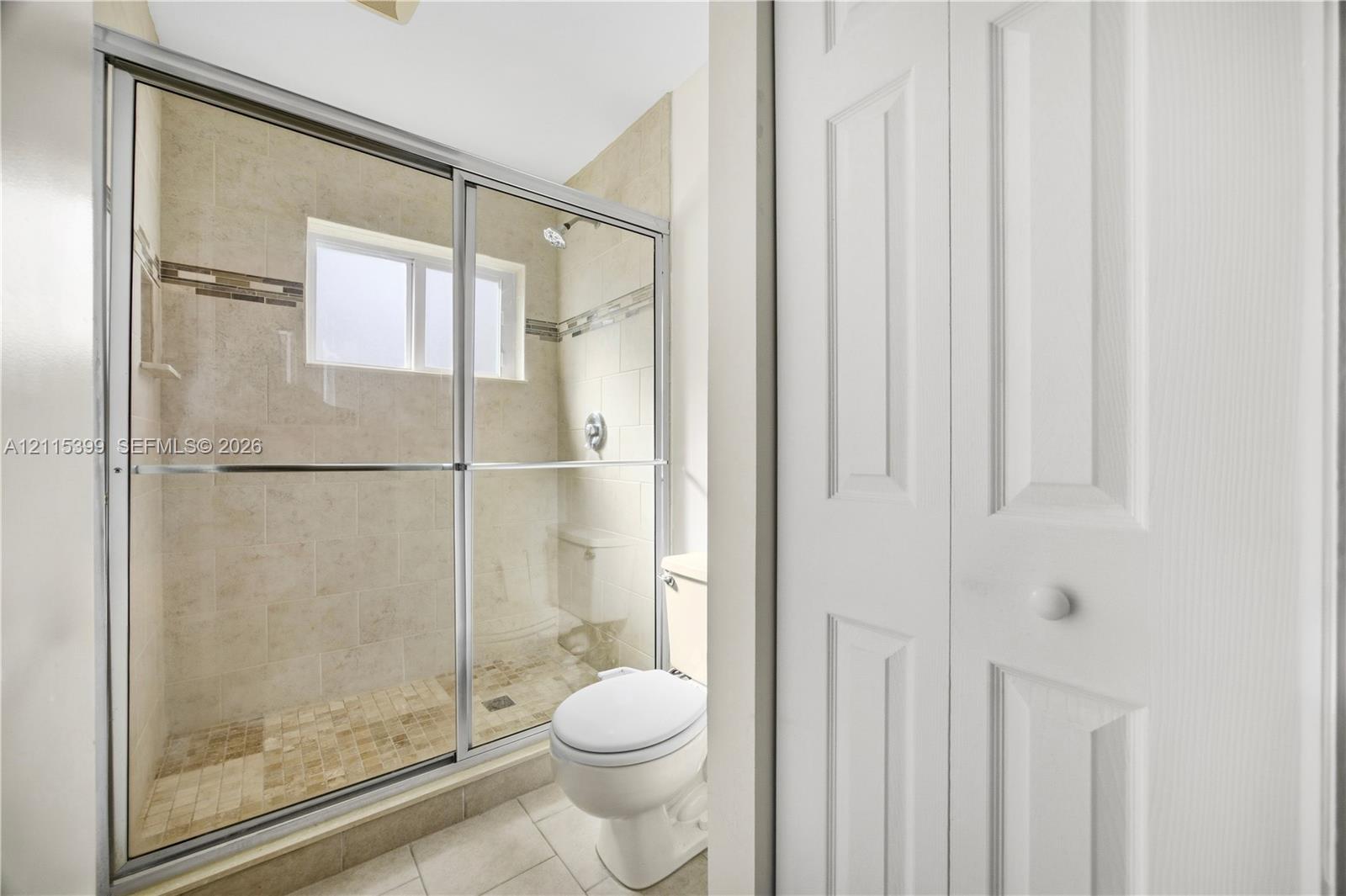 Shower enclosure with sliding glass doors, neutral tile wall surround with decorative inlay, inset window, and classic white paneled closet doors