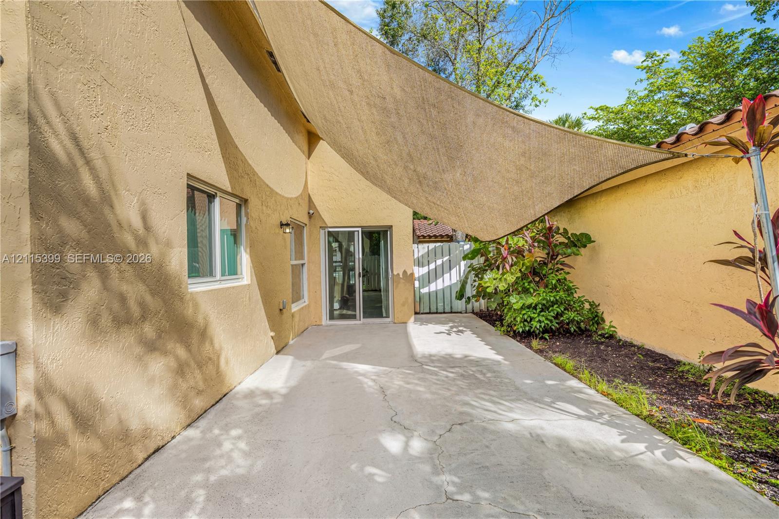 Concrete patio featuring a retractable shade sail, stucco exterior walls, sliding glass door, and integrated planting beds