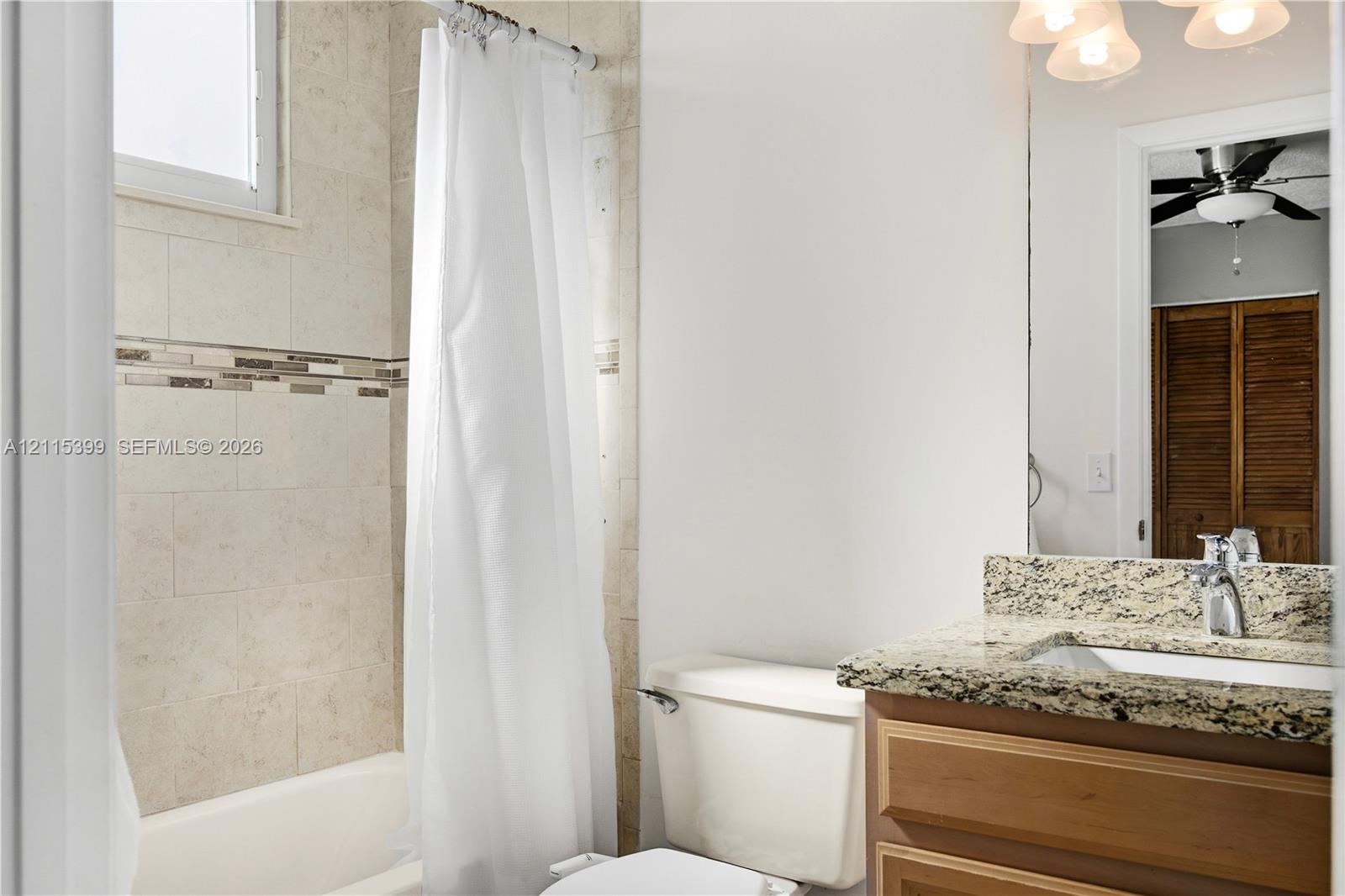 Bathroom featuring stone-finish vanity countertop with undermount sink, wood-finish cabinetry, neutral tile tub surround with decorative mosaic accent, and multi-light fixture