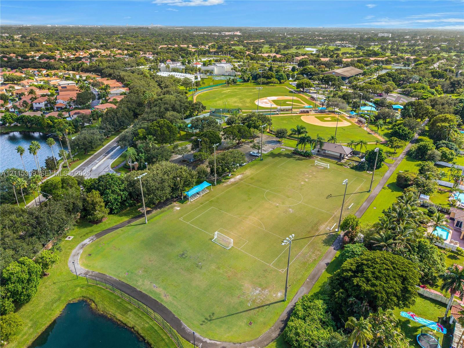 Aerial perspective featuring a multi-purpose grass field, baseball diamonds, perimeter walking path, and pond