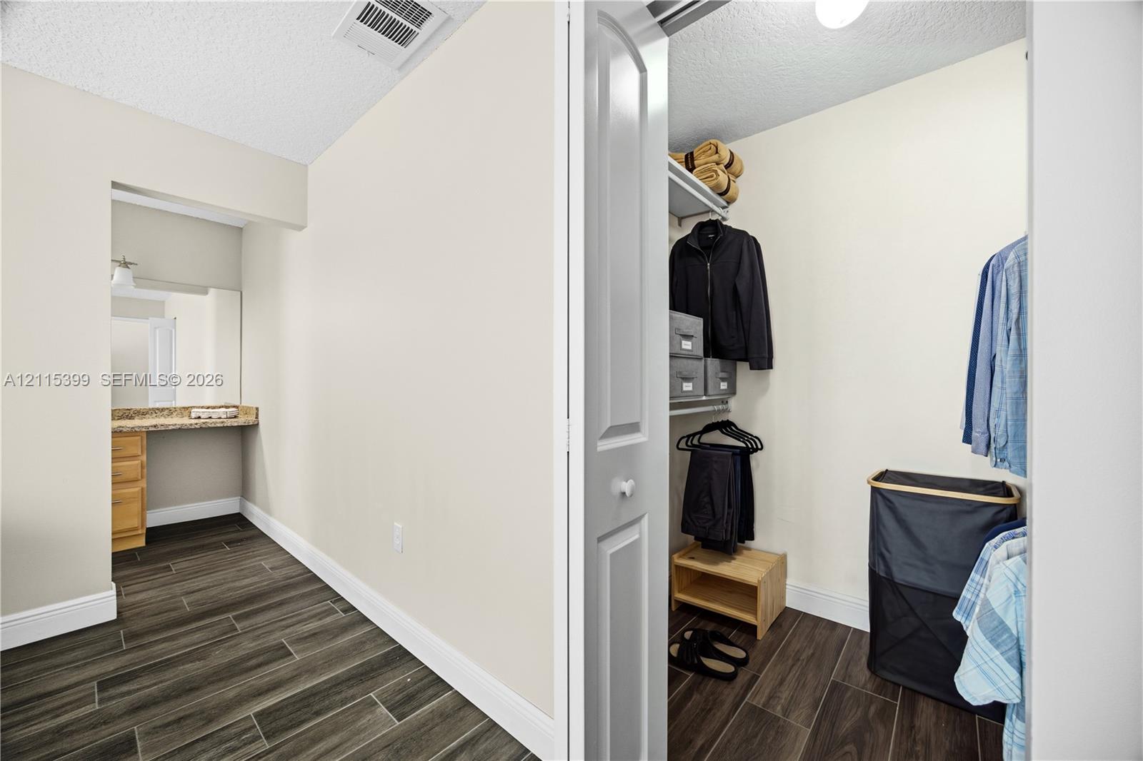 Walk-in closet featuring built-in wire shelving and wood-finish flooring, complemented by a vanity area with wood cabinetry and stone-finish countertop