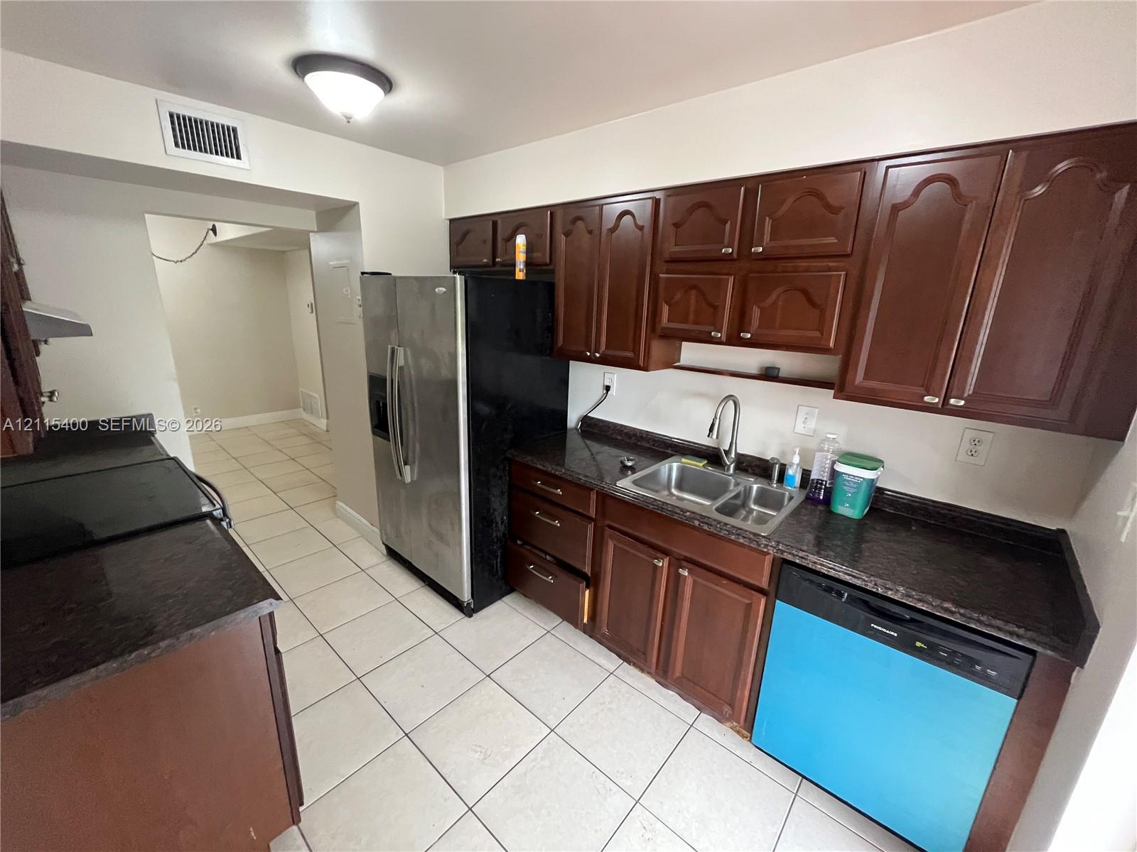 Kitchen featuring dark wood-finish cabinetry, stone-surface countertops, stainless steel refrigerator, dual-basin sink with arched faucet, and tiled flooring