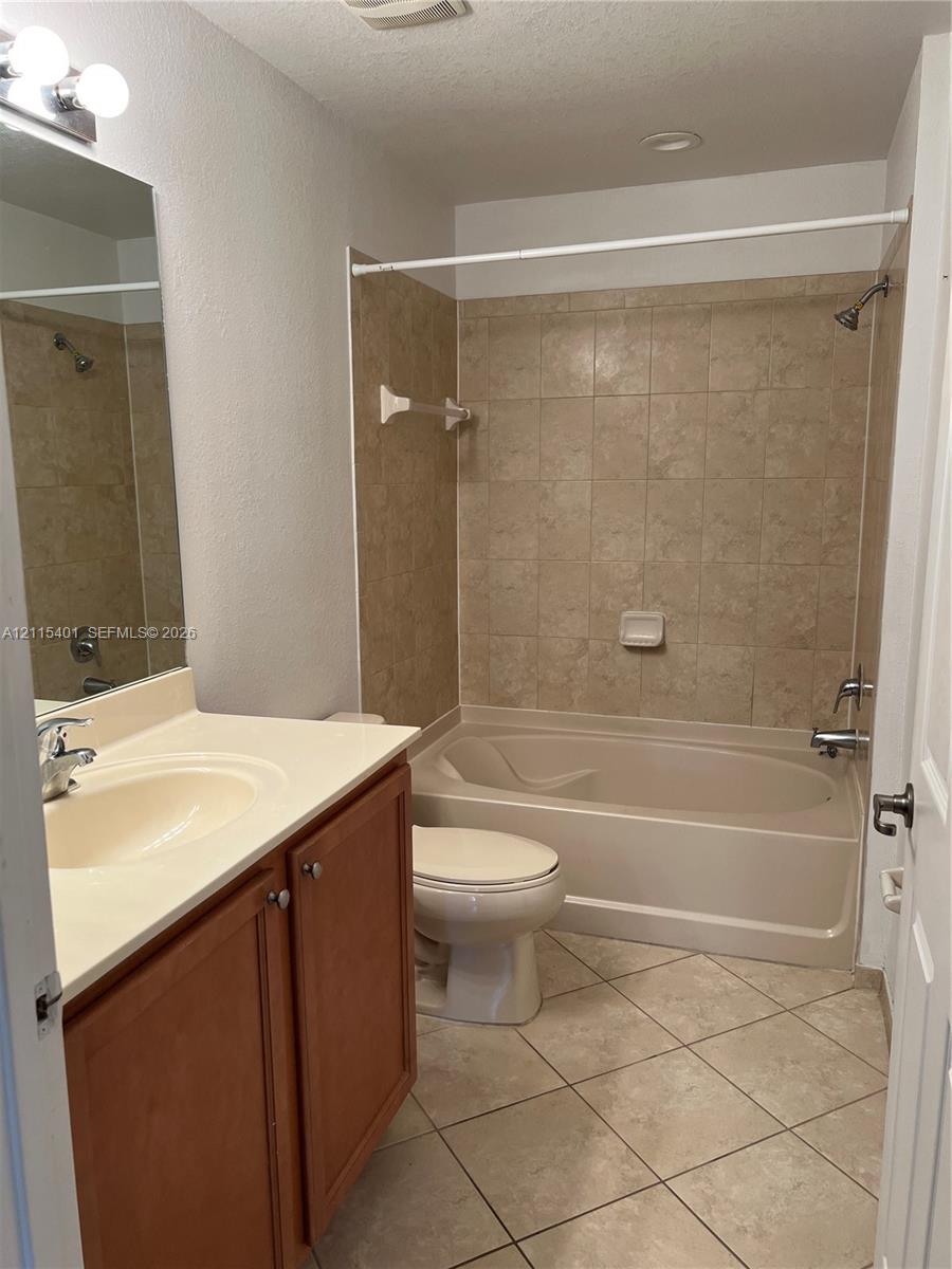 Bathroom featuring a vanity with a solid-surface countertop, ceramic tile flooring, a tub-shower combination with full-height tile surround, and a wall-mounted mirror with integrated lighting