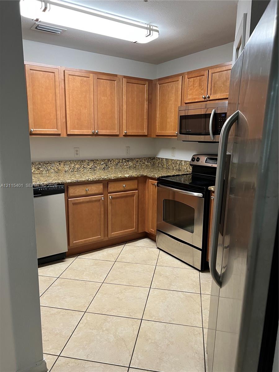 Kitchen featuring warm-tone wood cabinetry, stone countertops, stainless steel appliances, and tile flooring