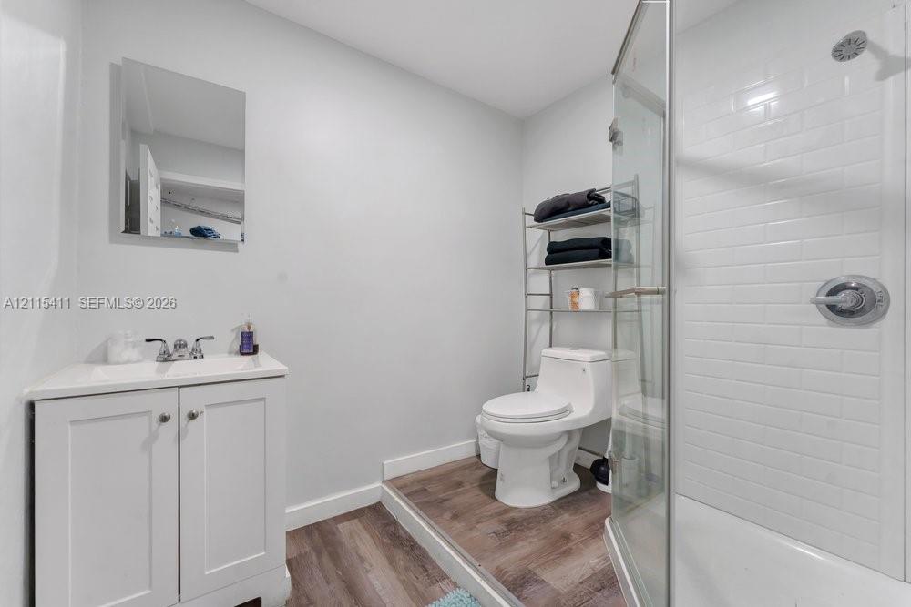 Bathroom featuring a white vanity cabinet with an integrated sink, wood-finish flooring, a toilet, and a glass-enclosed shower with white subway tile surround