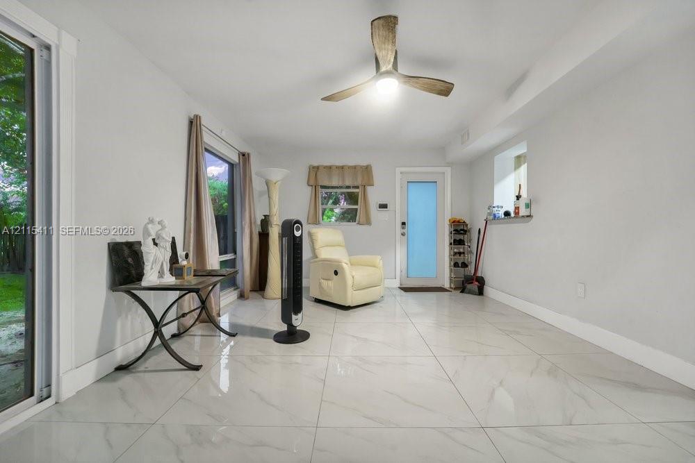 Large-format tile flooring with a neutral finish, multi-blade ceiling fan with integrated light, glass-paneled exterior door, and sliding glass door with external exposure