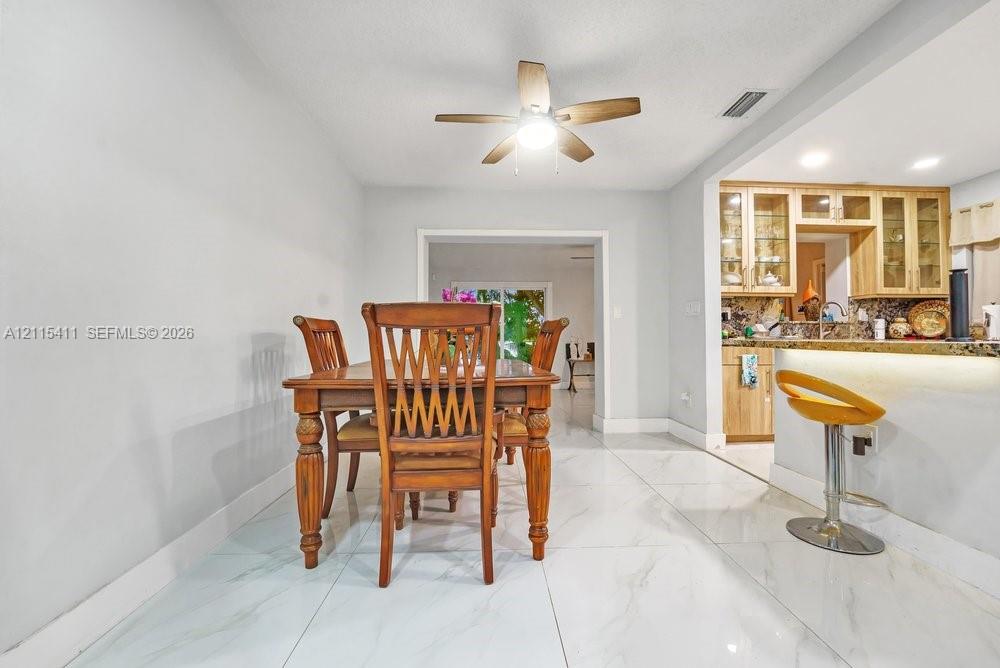 Dining area featuring large-format tile flooring, a ceiling-mounted fan with integrated lighting, and a kitchen pass-through with under-counter lighting and wood-finish cabinetry
