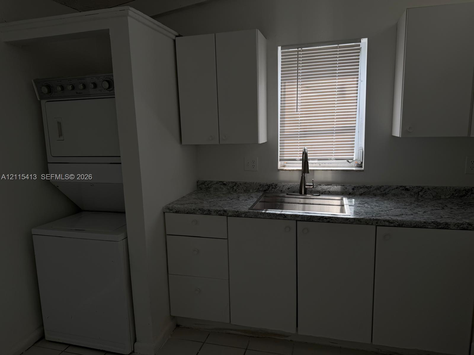 Utility space featuring white cabinetry, stone-finish countertops, stainless steel sink with a high-arch faucet, and an integrated stacked laundry unit