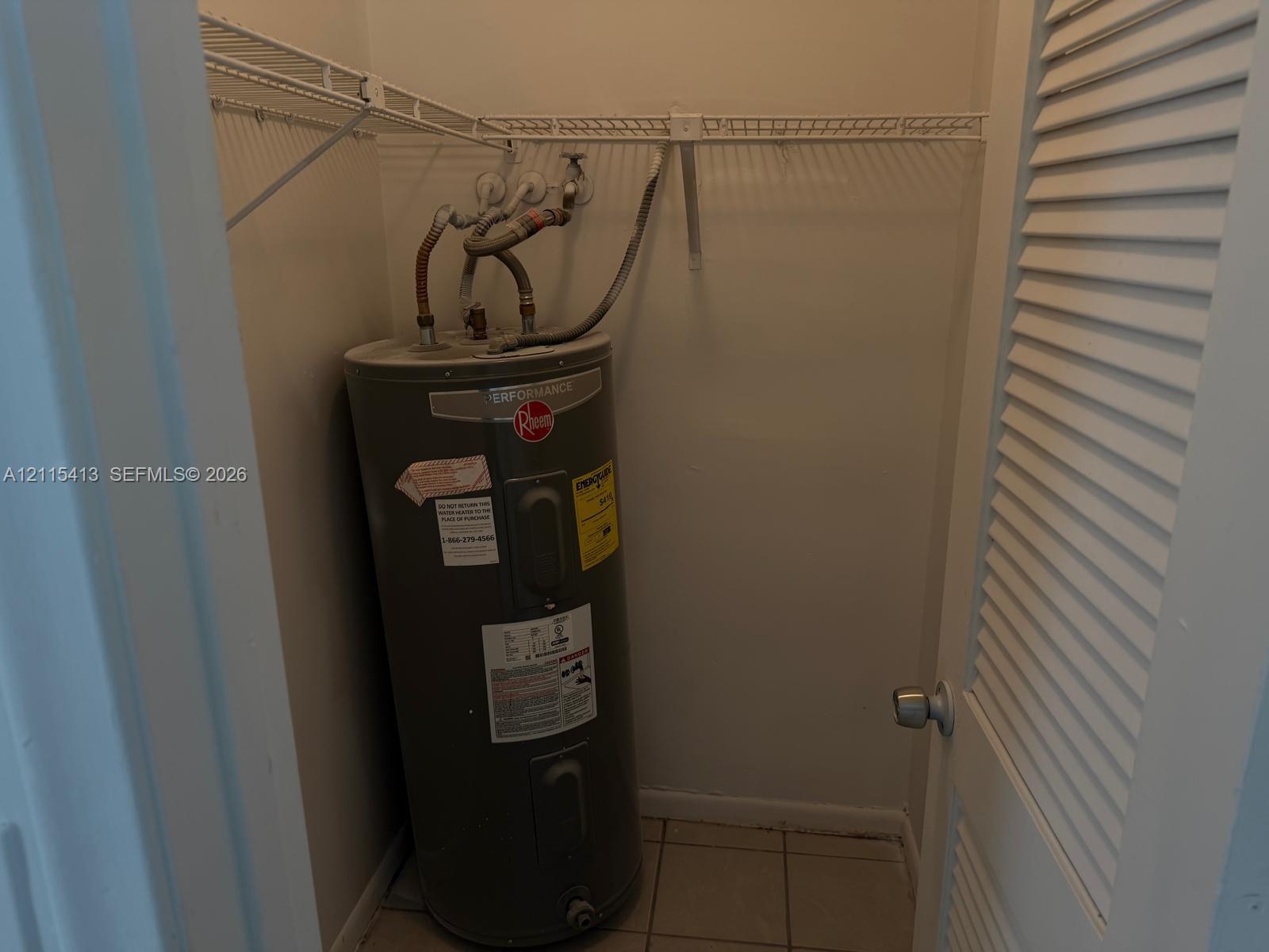 Utility closet featuring a water heater, tiled flooring, and overhead wire shelving behind a louvered door