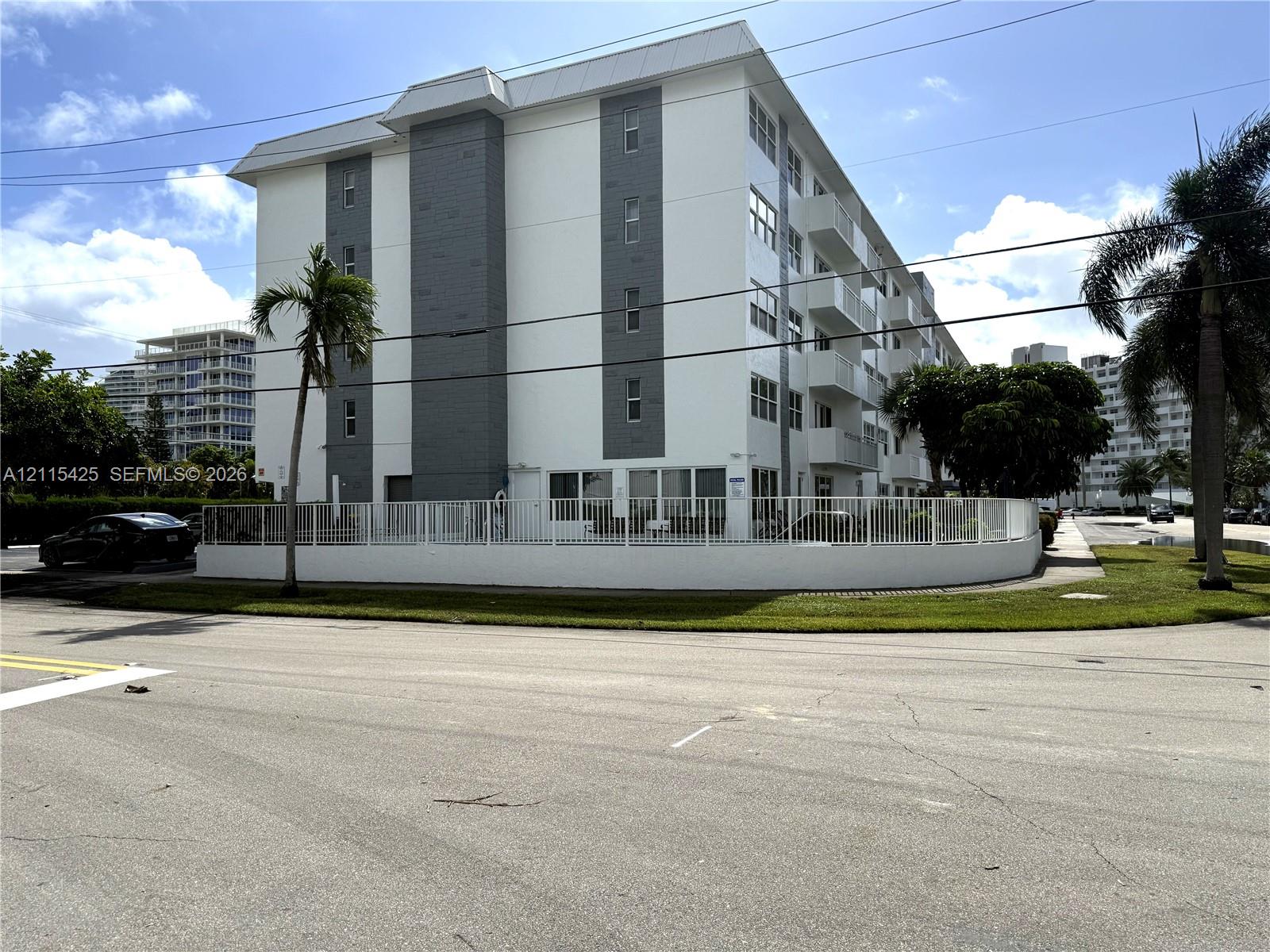 Multi-story building featuring white stucco exterior, grey paneled accents, private balconies, and a perimeter fence enclosing the property grounds