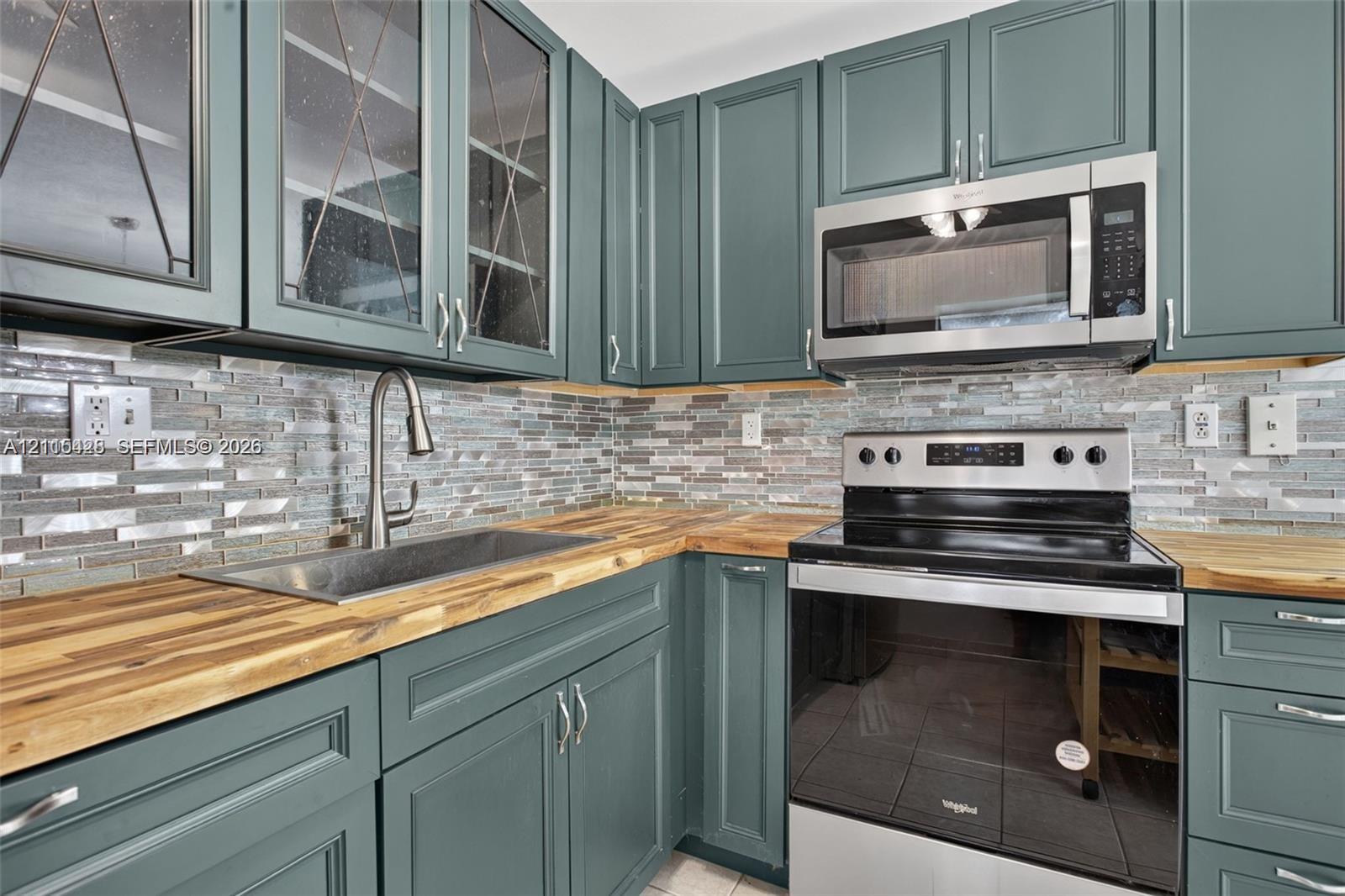 Teal cabinetry featuring glass-front uppers, butcher block countertops, stainless steel undermount sink with gooseneck faucet, horizontal glass mosaic tile backsplash, and stainless steel appliances